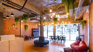 A modern interior with exposed brick walls, wooden floors, and hanging plants. There are seating areas with tables and chairs, and a reception desk. - Kaen Hostel (Pattaya)