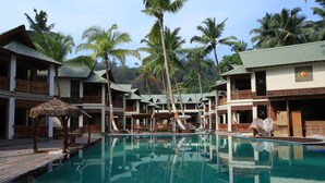 A pool area with multiple buildings, palm trees, and a thatched umbrella. - Sandyy Wavess Beach Resort (Port Blair)