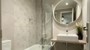 Un baño moderno con ducha envuelta en vidrio, inodoro blanco, bañera independiente y un espejo redondo sobre un lavamanos.