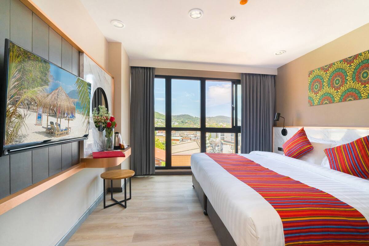 A hotel room with a bed, a TV, a window with a view, a small table, and a chair.