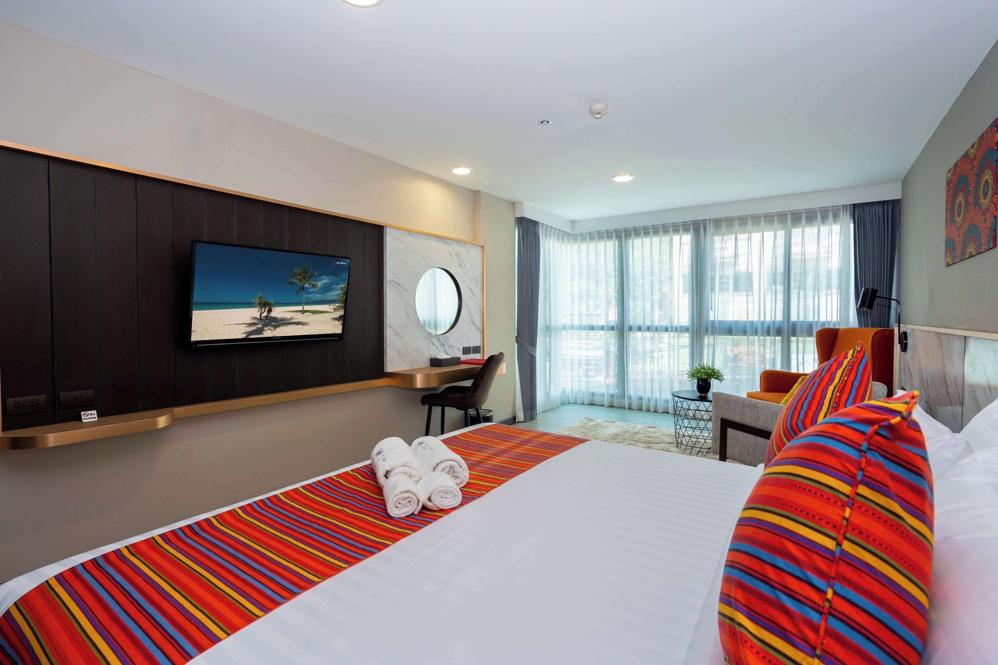 A hotel room with a large bed, a TV mounted on the wall, a desk with a chair, and a window with curtains.