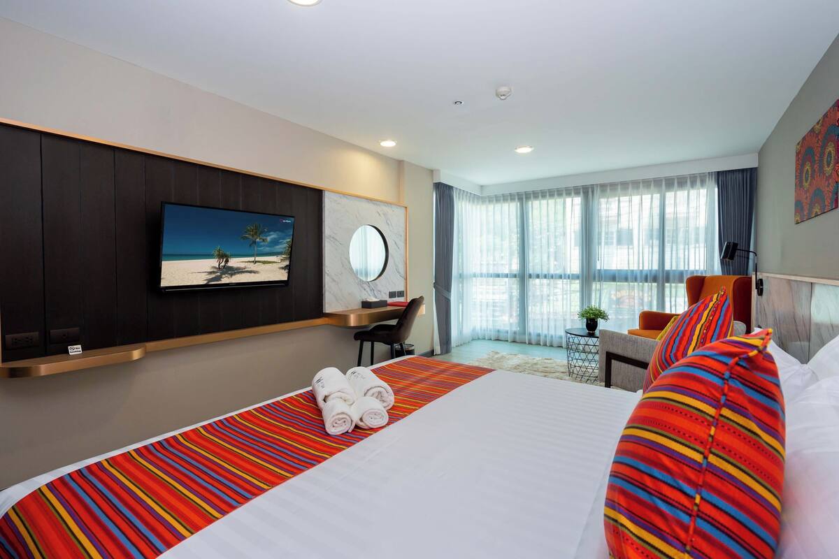 A hotel room with a large bed, a TV mounted on the wall, a desk with a chair, and a window with curtains.