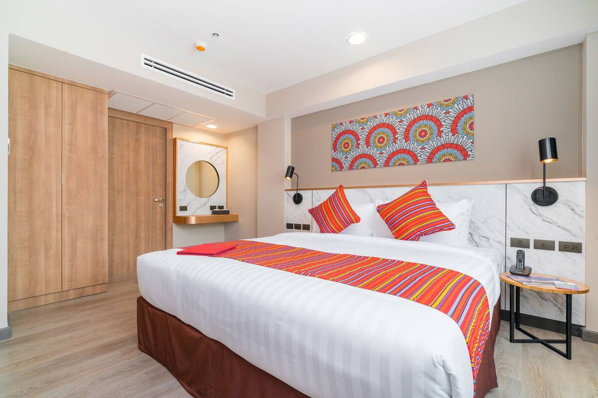 A hotel room with a large bed, wooden wardrobe, marble headboard, and a colorful wall art piece.