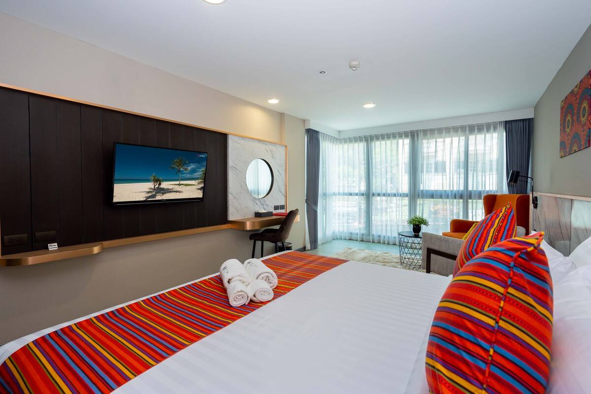 A hotel room with a large bed, a TV mounted on the wall, a desk with a chair, and a window with curtains.