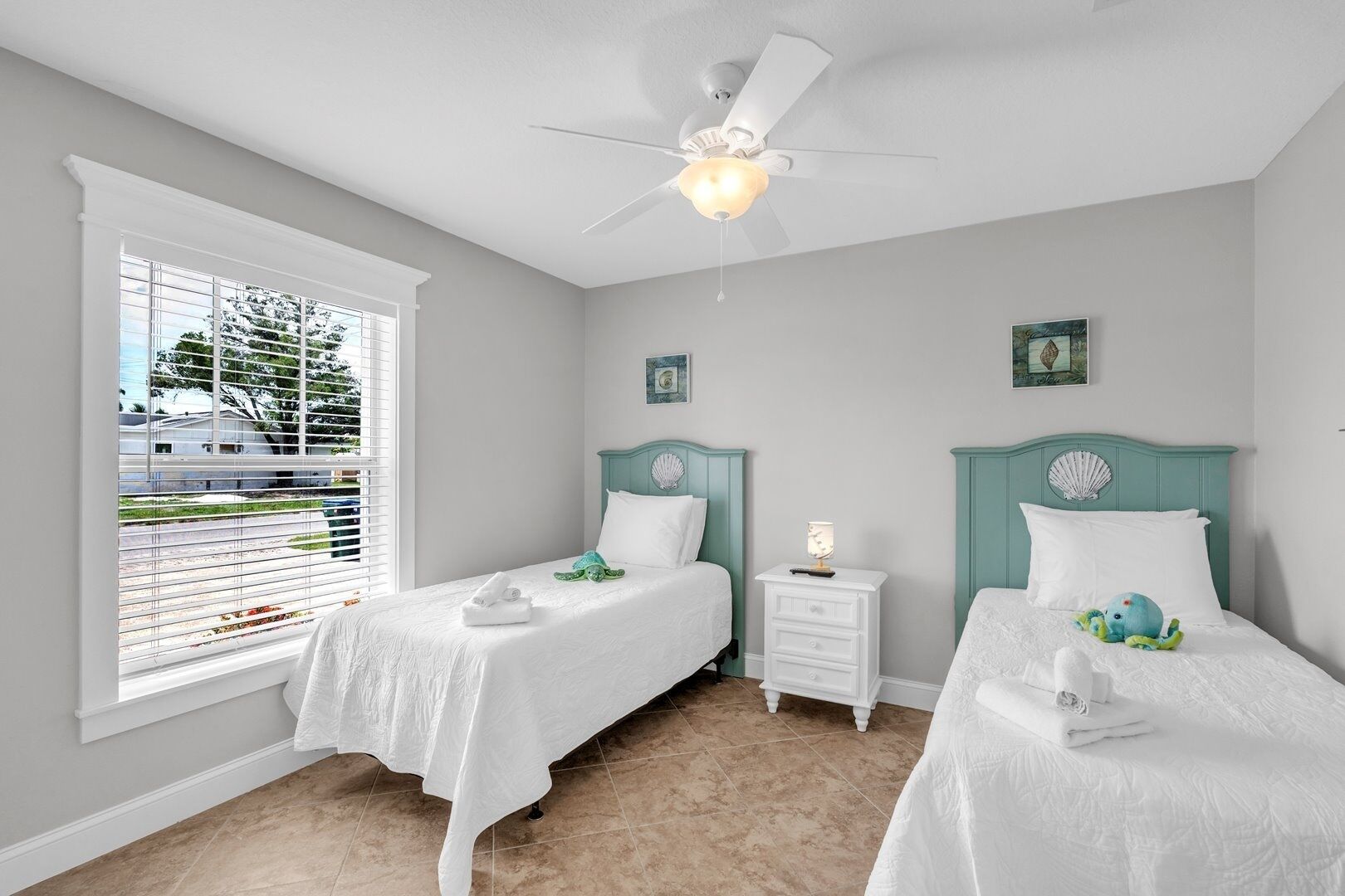 A bedroom with two single beds, a ceiling fan, a nightstand with a lamp, and a window with blinds.