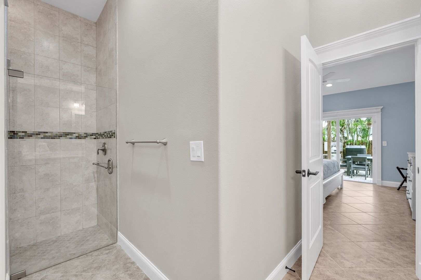 A bathroom with a shower, a towel rack, and a door leading to a bedroom with a bed and a table.
