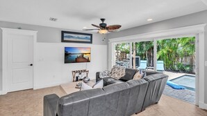 Amplia sala de estar con sofá de cuero gris, ventilador de techo y ventana grande que ofrece vista a un patio trasero con piscina.