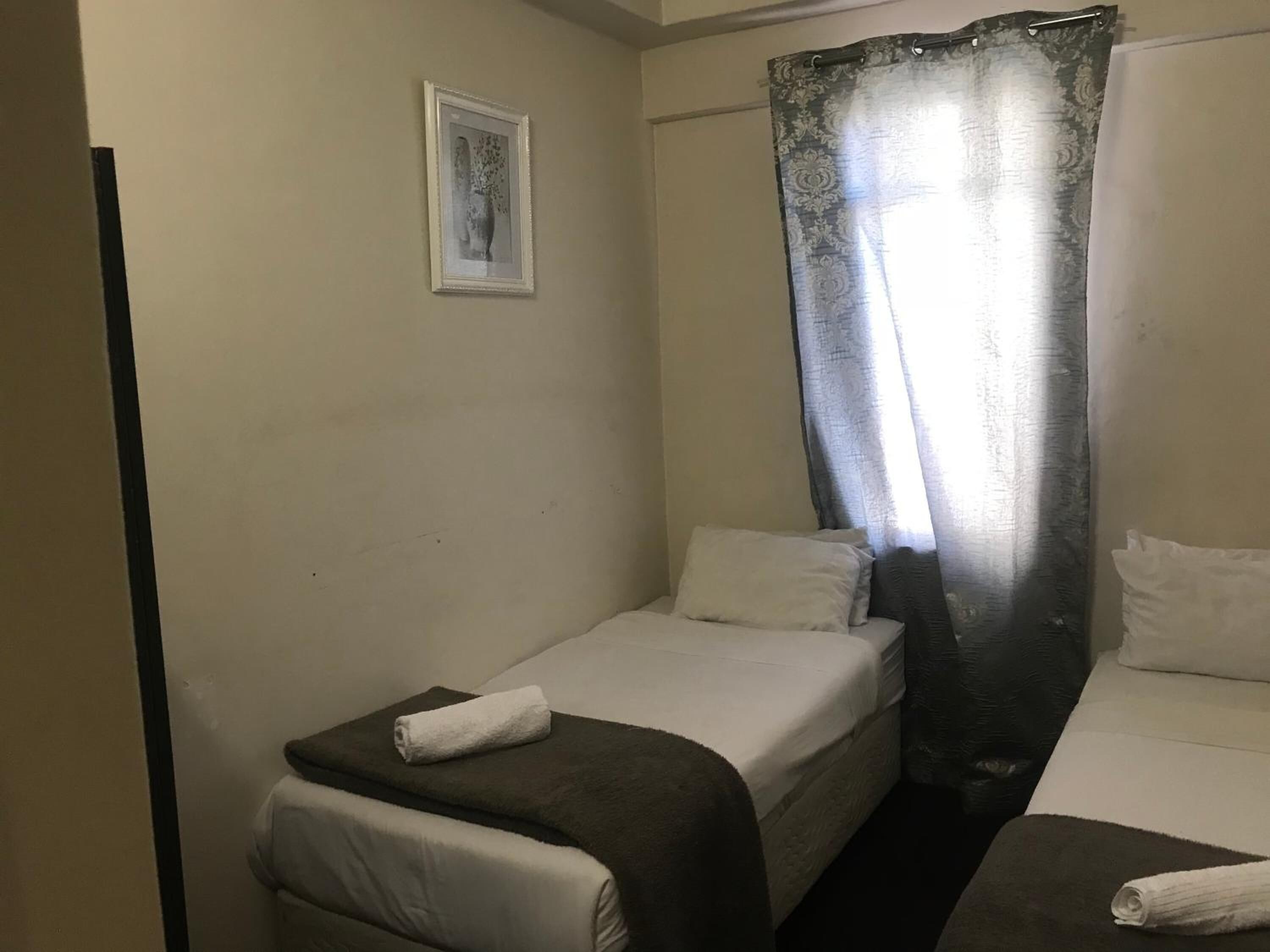 Une petite chambre avec un lit simple, recouverte d’une couvre-lit de couleur sombre, un oreiller blanc et une serviette enroulée posée sur le lit. Un tableau encadré est accroché au mur et un rideau est partiellement tiré sur le côté.