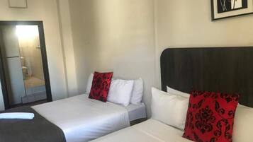A hotel room with two beds, each with white bedding and red patterned pillows. There is a dark headboard on the right side of the beds. A framed picture of a red rose is visible on the wall above the beds.