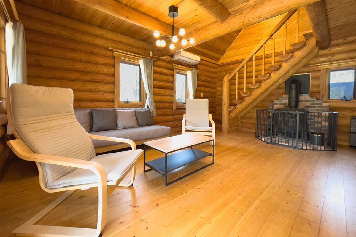 Interior de una cabaña de troncos con un sofá, dos sillones, una mesa de centro, una chimenea y una escalera.