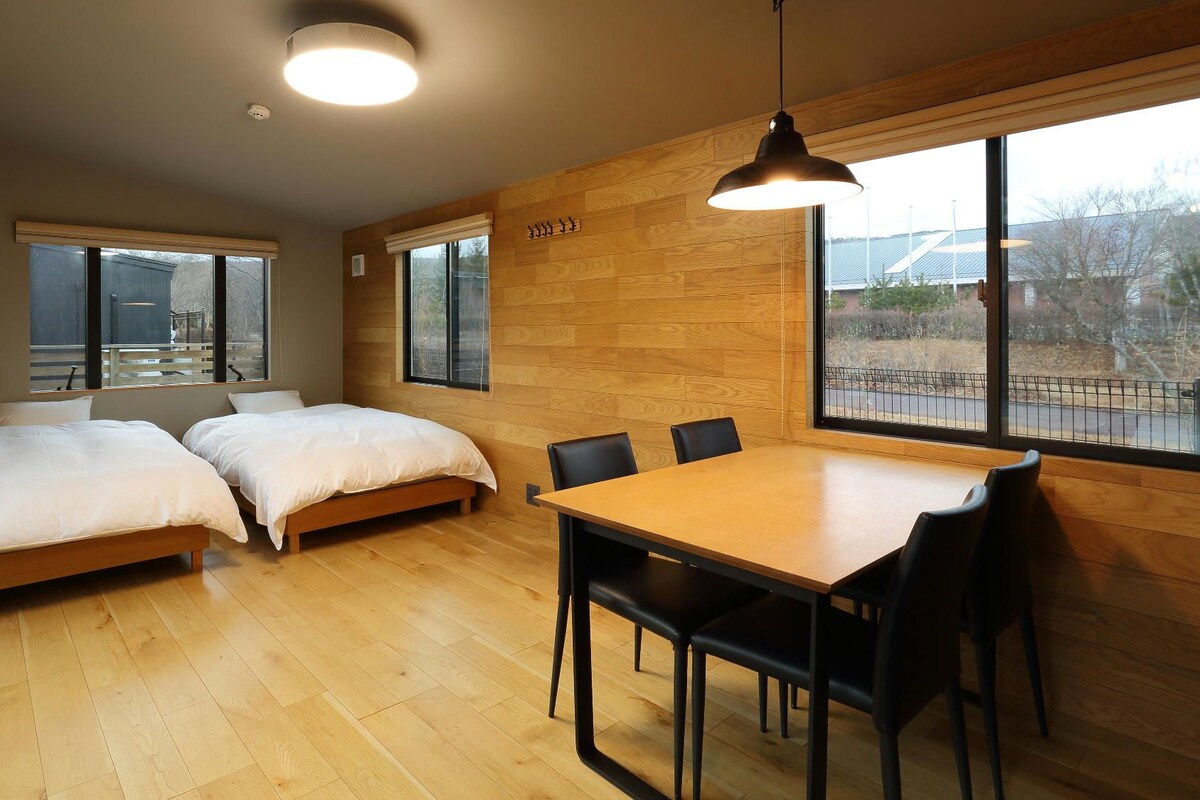 Una habitación con dos camas, una mesa de comedor con sillas, ventanas amplias y una pared de madera.
