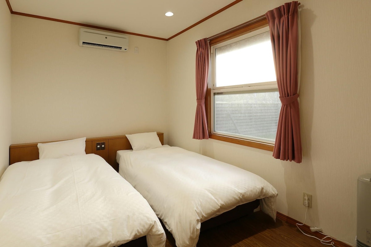 Habitación de hotel con dos camas, un equipo de aire acondicionado, ventana con cortinas y suelo de madera.