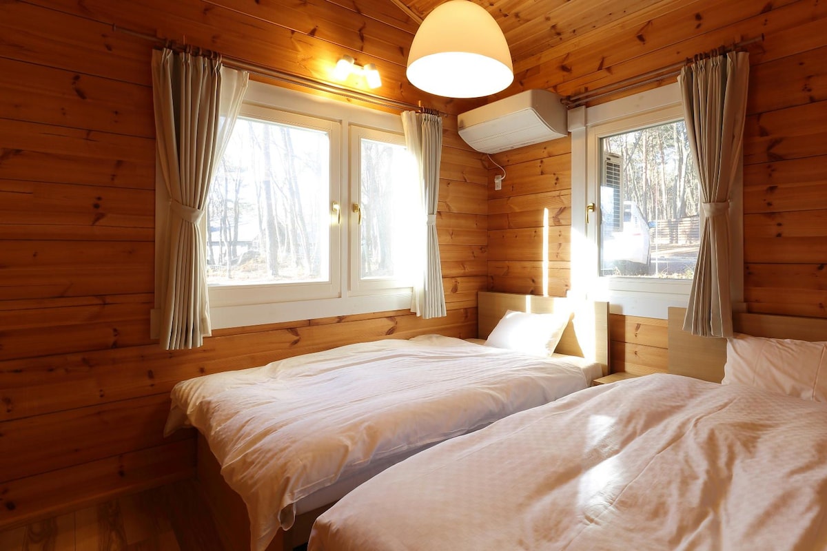 Habitación de cabaña de madera con dos camas, una lámpara de techo y ventanas con cortinas.