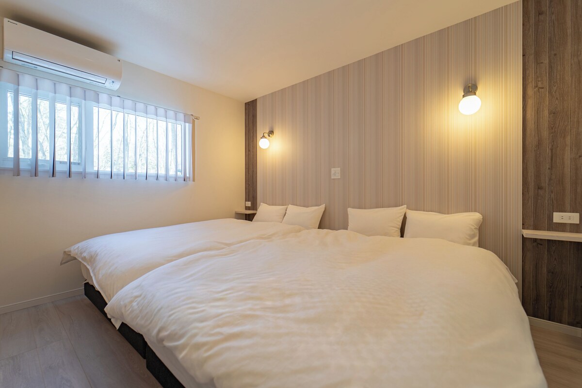 Una cama grande en una habitación con ventana, una lámpara de pared y un panel de madera.