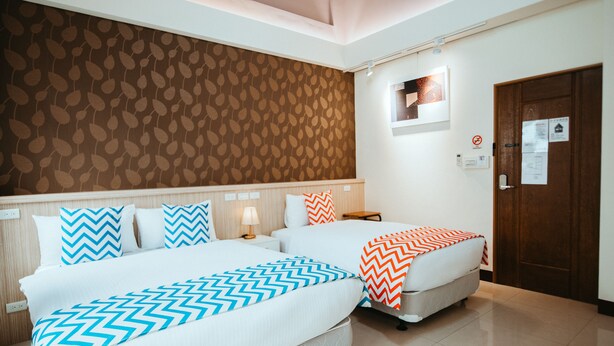 Ein Hotelzimmer mit zwei Betten, einer gemusterten Wand, einer Holztür und einem kleinen Tisch mit einer Lampe.