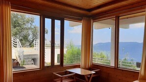 A room with a bed, a table, and chairs. There are large windows with curtains, and a view of greenery and mountains. - Snow View Luxury Homes (Dhari)