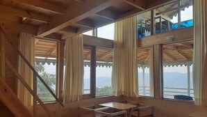 A wooden cabin room with a dining table and chairs, large windows with curtains, and a view of the outdoors. - Snow View Luxury Homes (Dhari)
