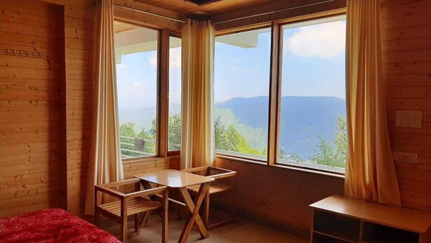 A room with a bed, a desk, and a chair, large windows with curtains, and a view of the outdoors. - Snow View Luxury Homes (Dhari)