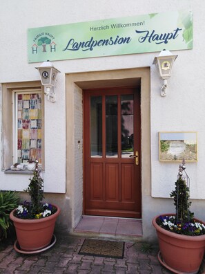 L’entrée d’un immeuble est agrémentée d’une enseigne indiquant « Landpension Haupt » et d’un message de bienvenue. Deux grands pots de fleurs sont placés de part et d’autre de la porte.