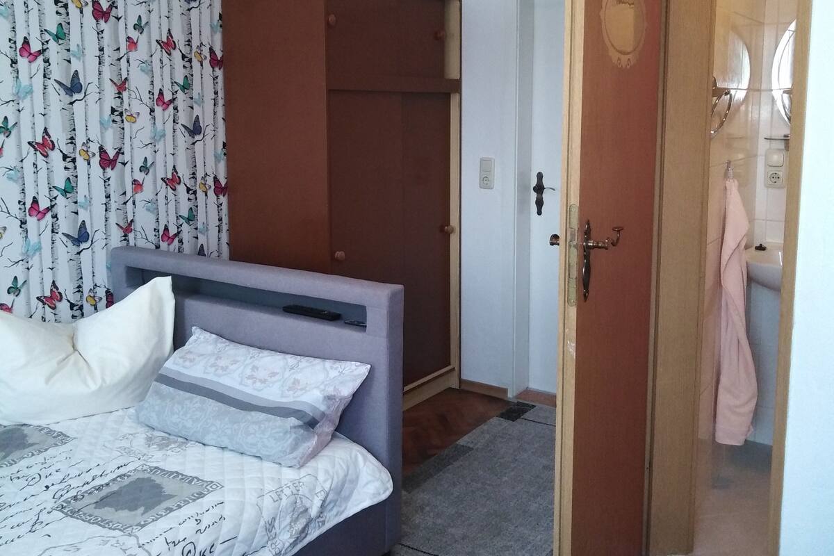 A bedroom with a bed, a wardrobe, a door leading to a bathroom, and a patterned curtain.