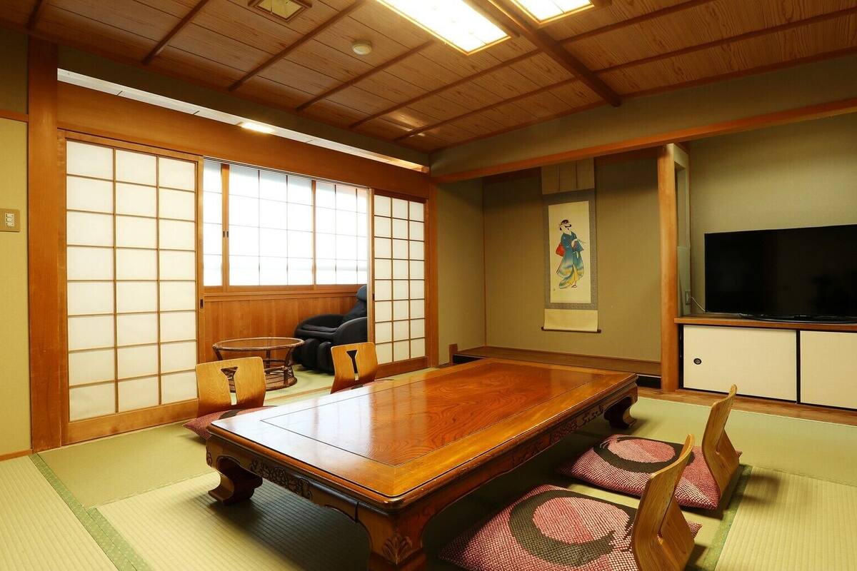 A traditional Japanese-style room with tatami flooring, a wooden dining table, tatami mats, a black leather sofa, a television, and sliding doors.