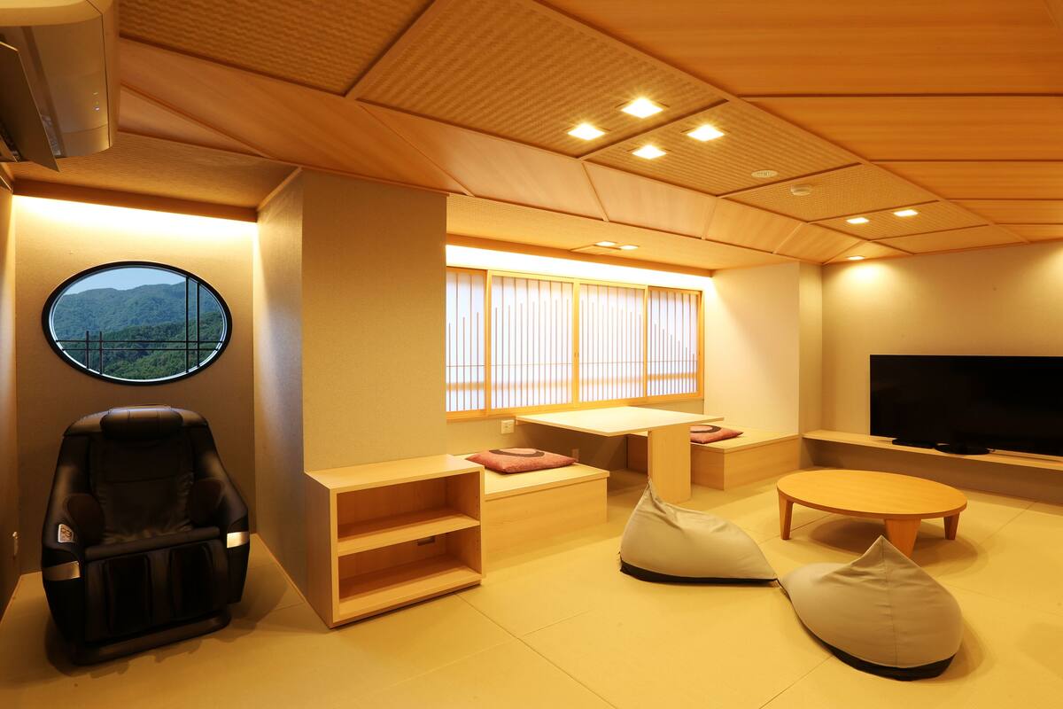 A modern room with a massage chair, a round window with a view, a TV, and a wooden ceiling.