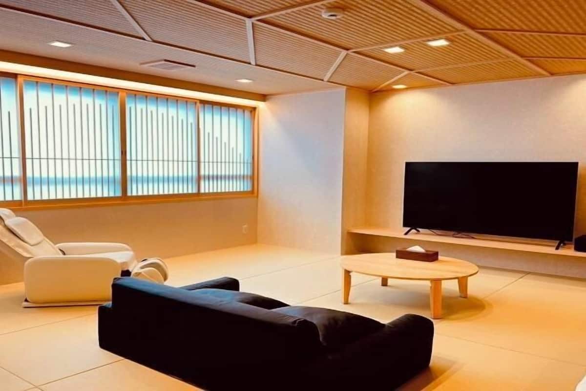 A modern living room with a flat-screen TV, a sofa, a coffee table, and a large window with blinds.