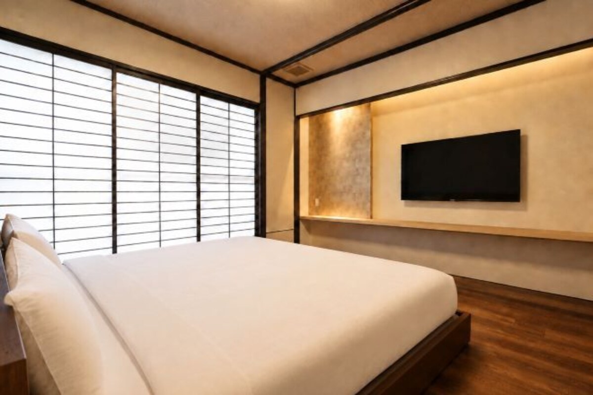 A bedroom with a bed, a television mounted on the wall, and a window with blinds.