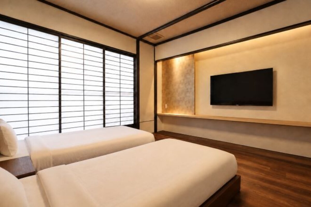 A hotel room with a traditional Japanese aesthetic, featuring tatami flooring, shoji screens, and a flat-screen TV mounted on the wall.