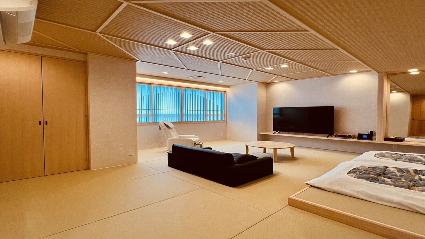 A modern room with a large bed, a TV, a massage chair, and a large window.