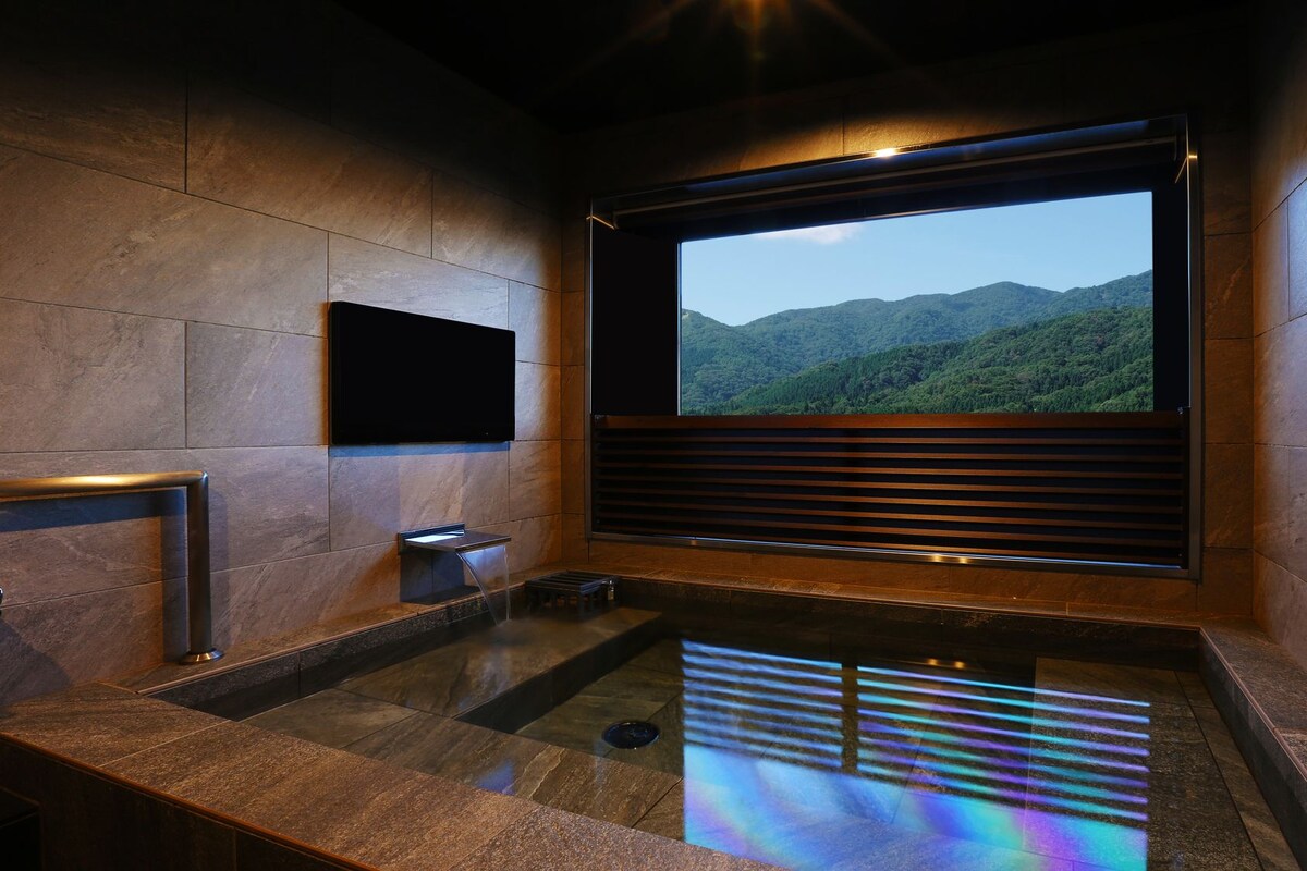 A modern bathroom with a large window, a flat-screen TV, and a reflective floor.