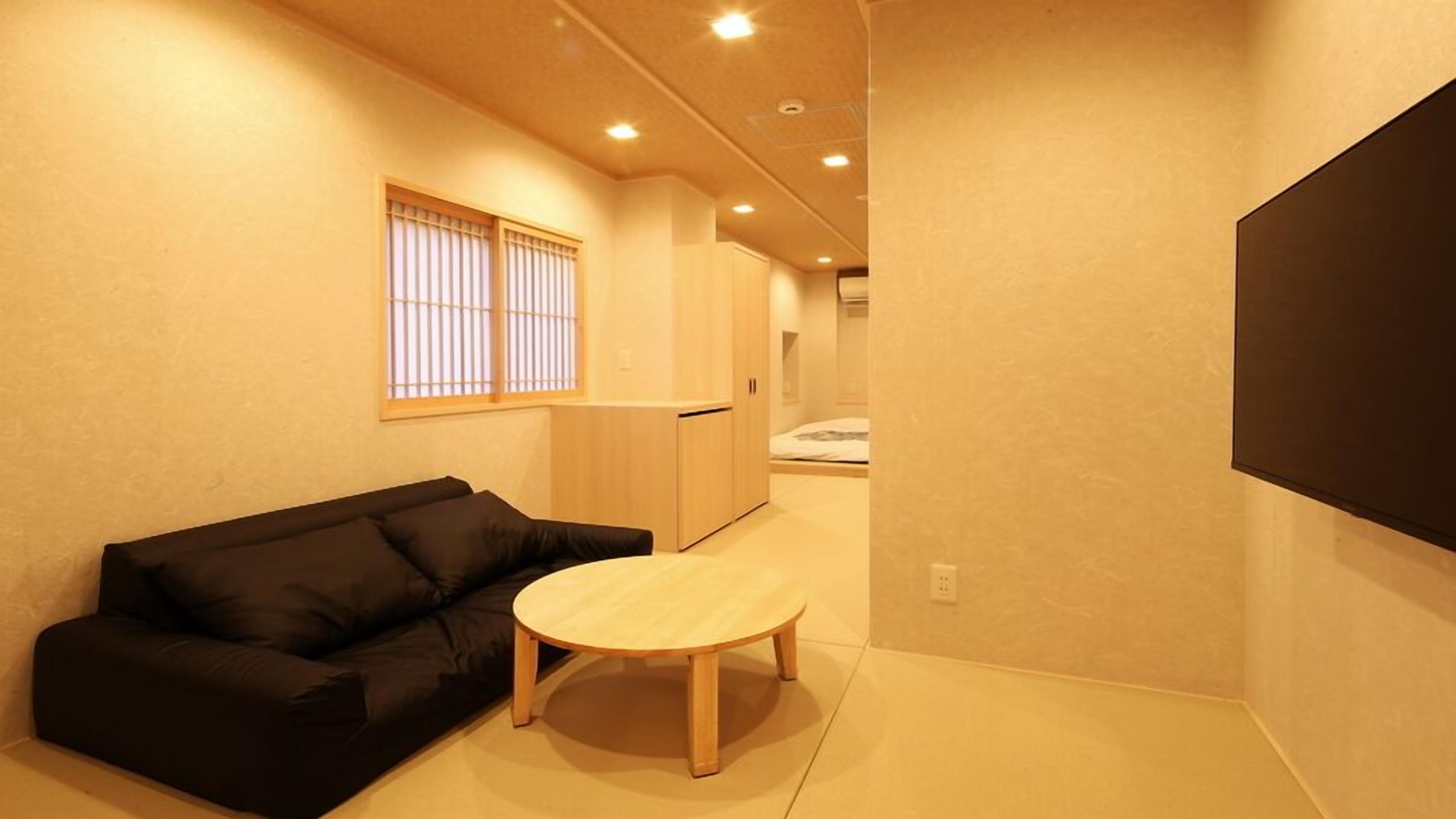 A room with a black sofa, a round wooden table, a window with vertical blinds, and a bed in the background.