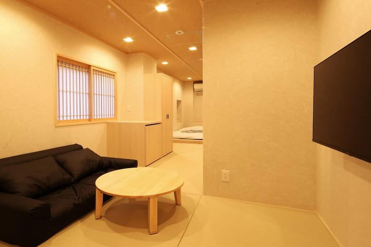 A room with a black sofa, a round wooden table, a window with vertical blinds, and a bed in the background.