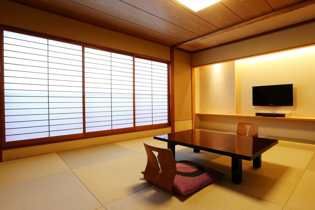 A traditional Japanese room with tatami flooring, a low table, chairs, a television, and sliding shoji doors.