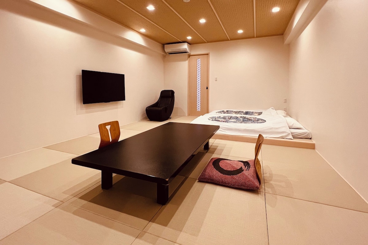 A room with a bed, a TV mounted on the wall, a black chair, a wooden chair, and a small table.