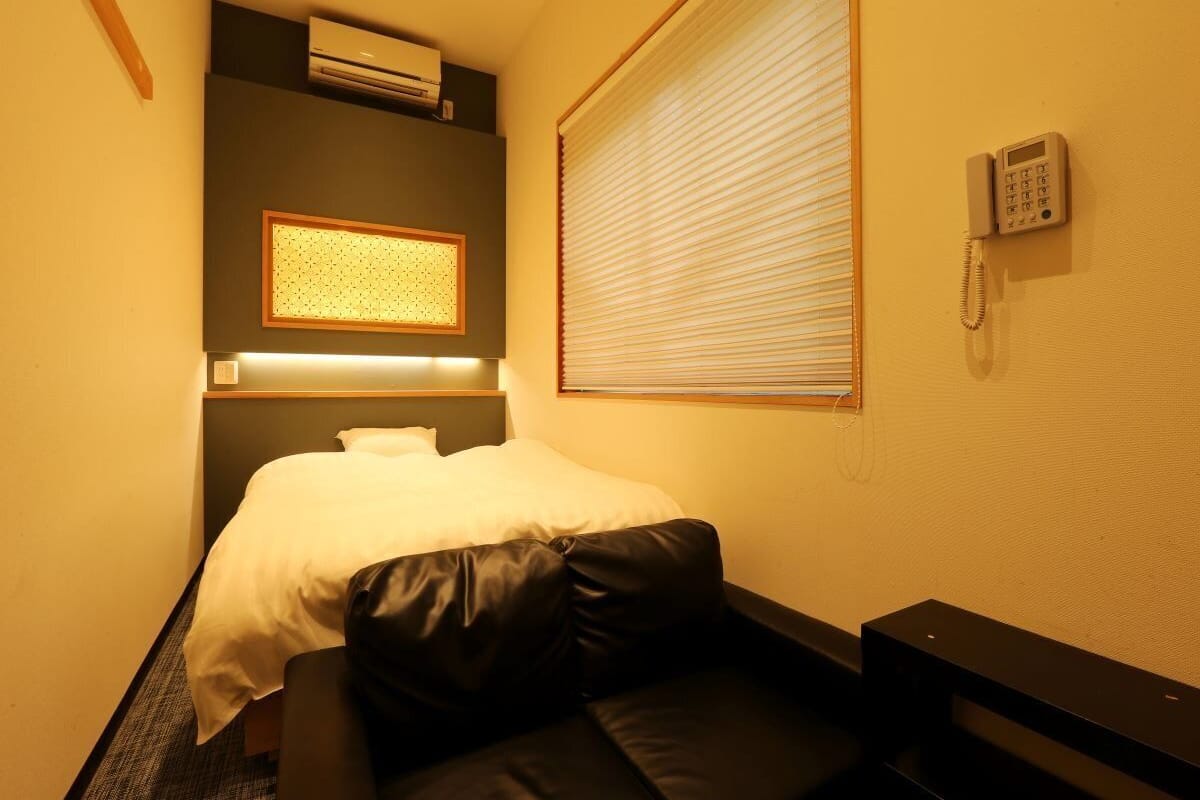 A compact hotel room with a bed, a small seating area, an air conditioning unit, a window with blinds, and a telephone.