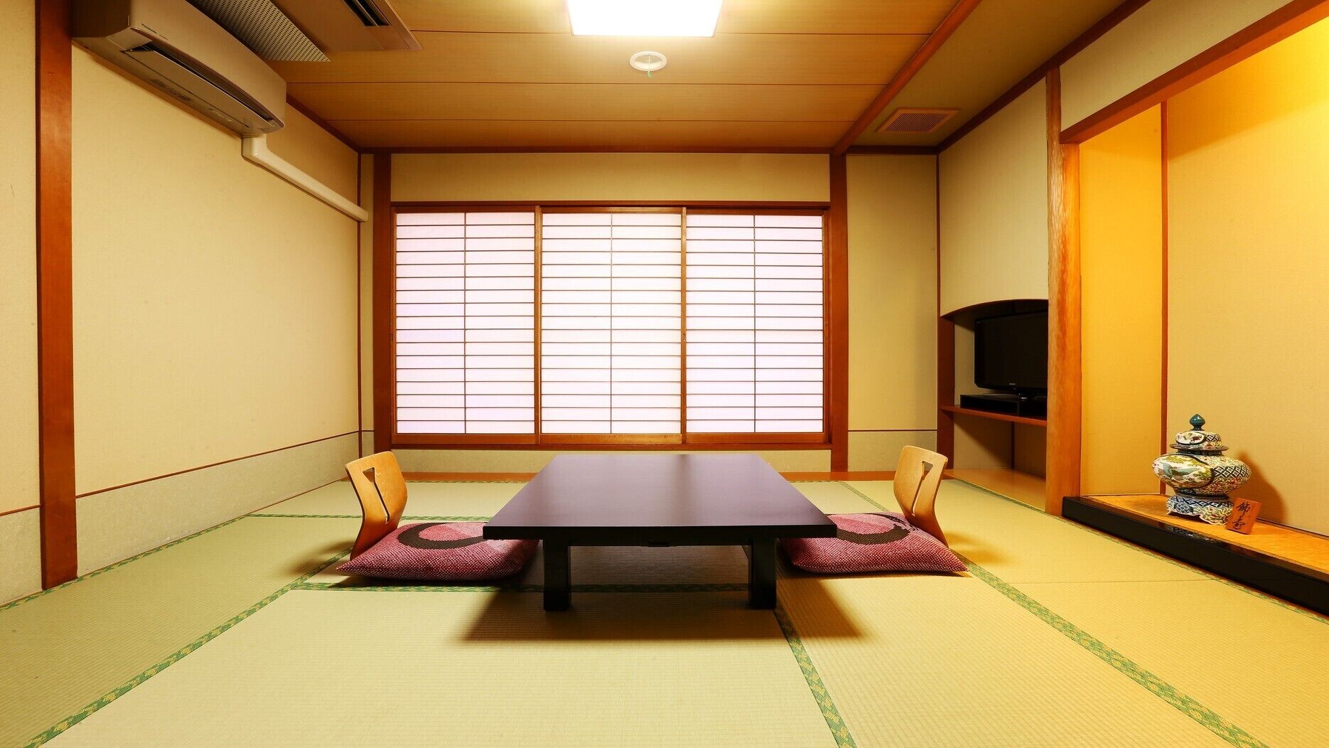 A traditional Japanese room with tatami flooring, a low table, two chairs, a sliding shoji screen, a television, and a decorative stack of pottery.