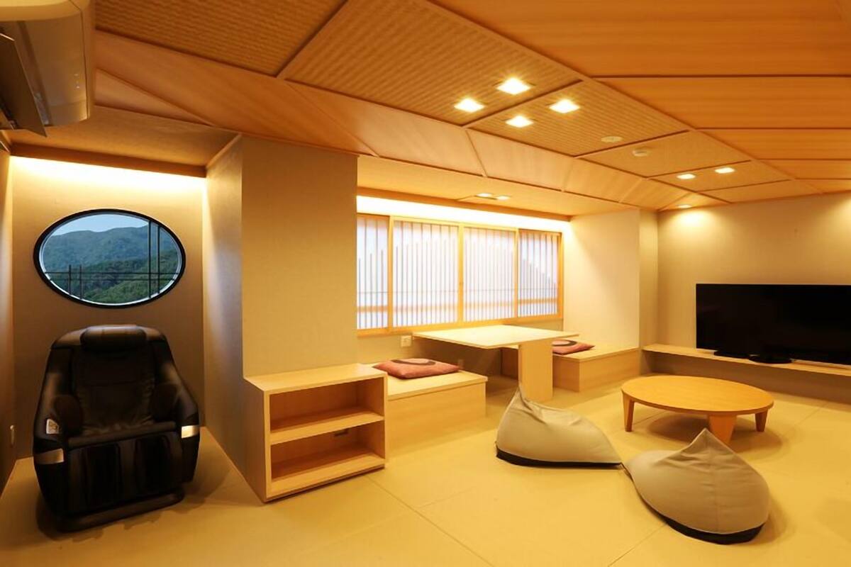 A modern living room with a sofa, ottoman, TV, and a window with blinds.