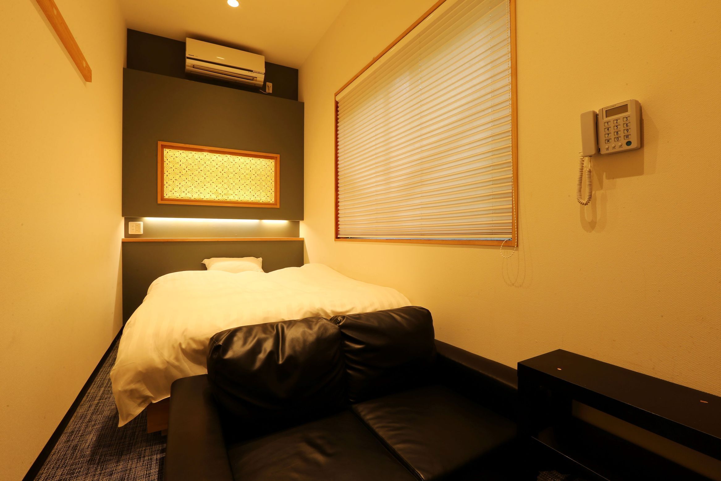 A hotel room with a bed, a small sofa, an air conditioning unit, a telephone, and a window with blinds.