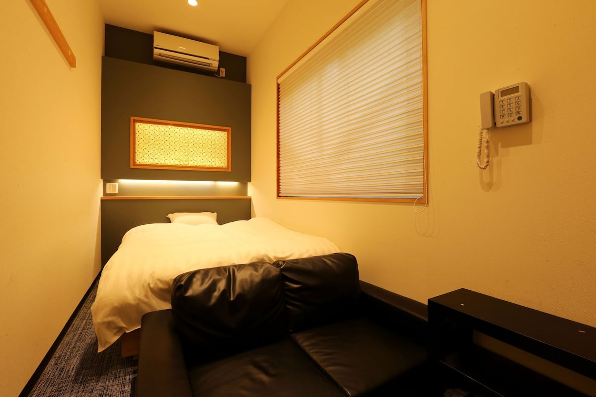 A hotel room with a bed, a small sofa, an air conditioning unit, a telephone, and a window with blinds.