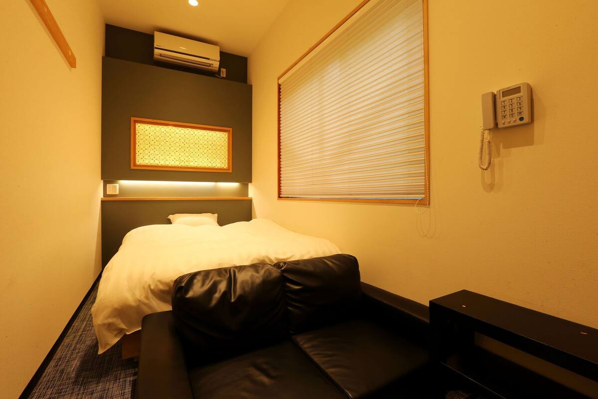 Ett hotellrum med en säng, en liten soffa, en luftkonditioneringsanläggning, en telefon och ett fönster med persienner.