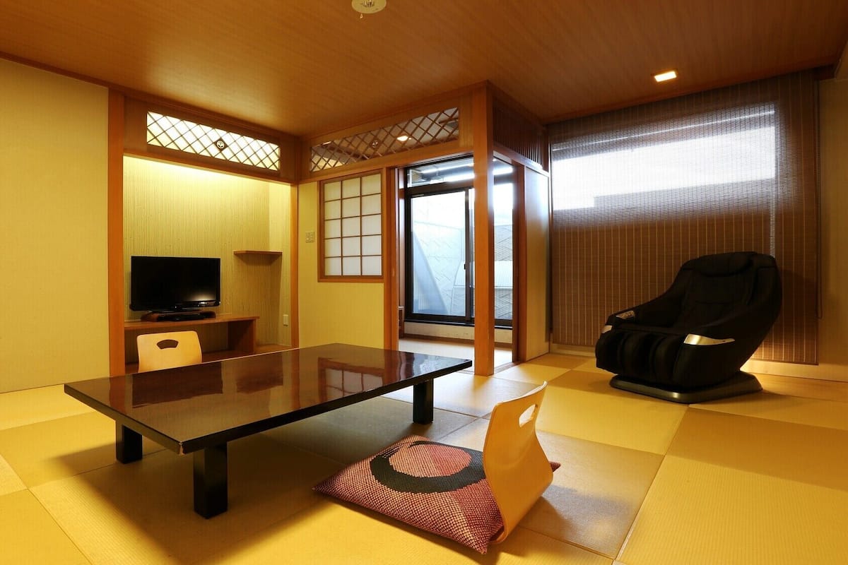 A traditional Japanese-style room with tatami flooring, a low table, a television, a massage chair, and sliding doors.