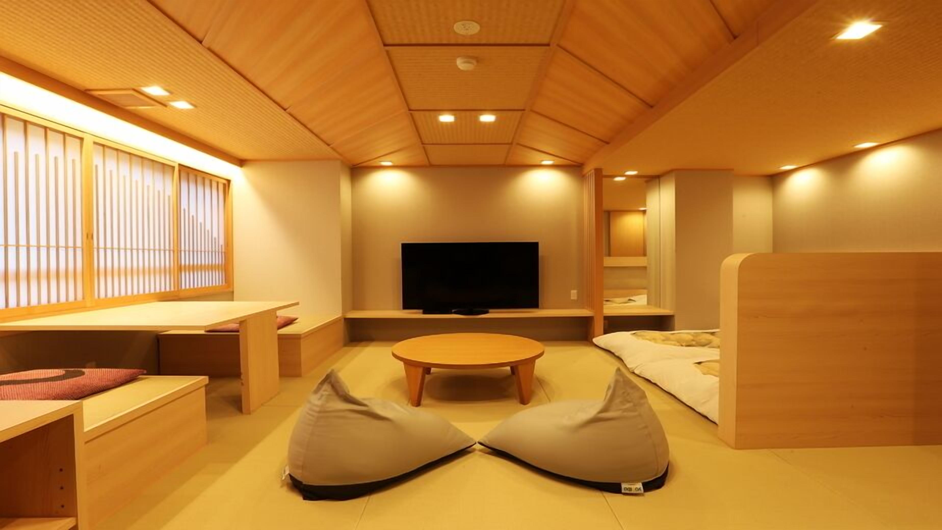 A modern room with a wooden ceiling, a flat-screen TV, a round table, bean bags, and a window with vertical blinds.