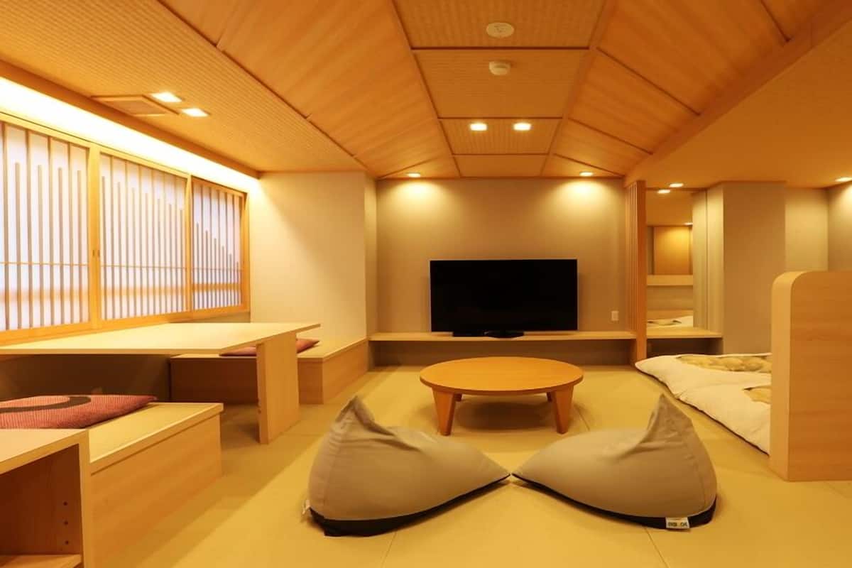 A modern room with a wooden ceiling, a flat-screen TV, a round table, bean bags, and a window with vertical blinds.