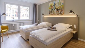 A hotel room with two single beds, a window with a view of buildings, a painting on the wall, and a desk with a chair. - Glück Auf Appartements Am Stadtpark (Duisburg)