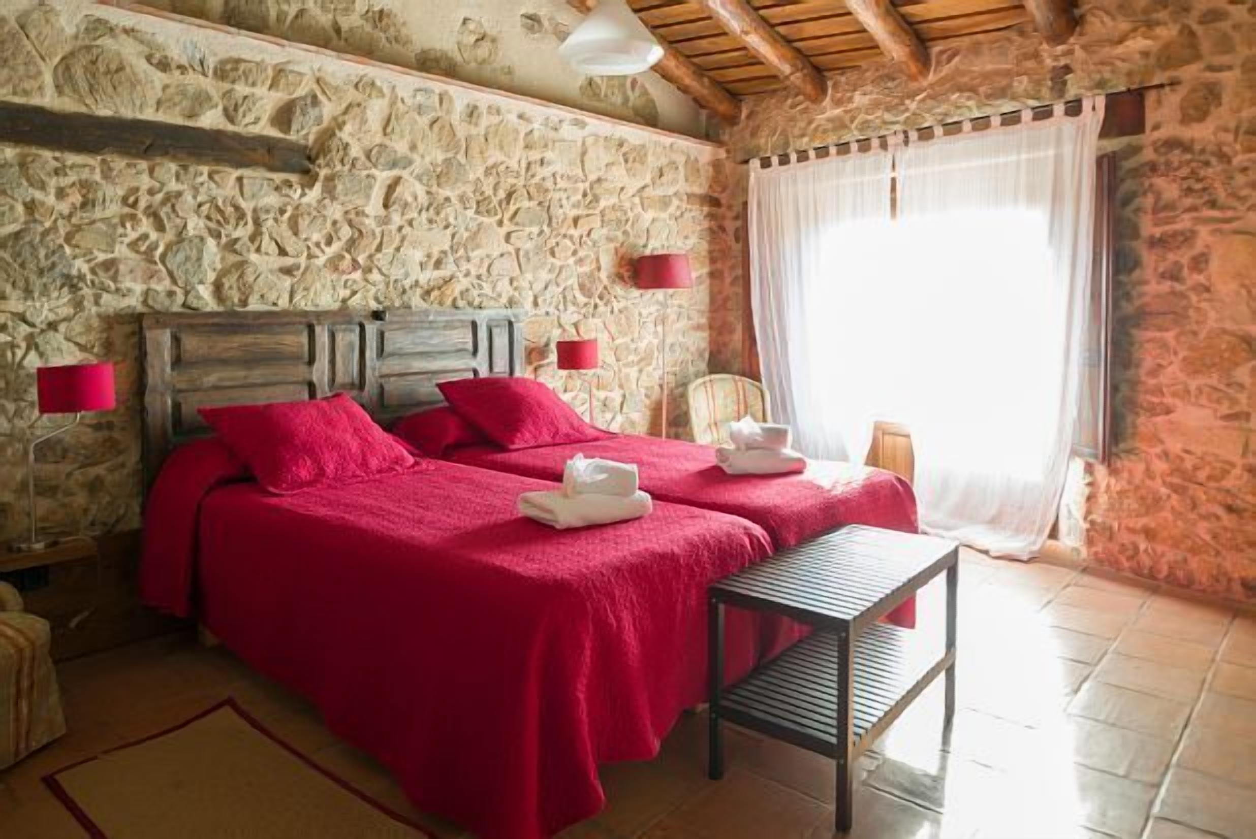 Una habitación con dos camas, paredes de piedra, techo de madera y una ventana con cortinas.