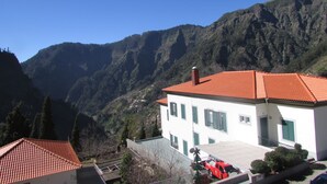 Ein Gebäude mit rotem Ziegeldach, weißen Wänden und grünen Fensterläden, eingebettet in eine gebirgige Landschaft.