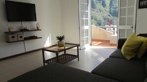 Ein Wohnzimmer mit einer Couch, einem Couchtisch und einem Flachbildfernseher. Eine Glastür führt auf einen Balkon mit Blick ins Grüne.
