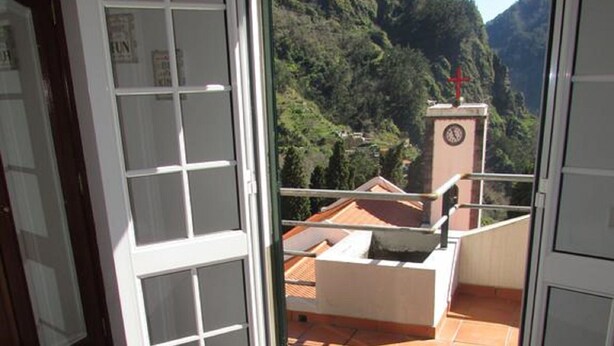 Ein Hotelzimmer mit Bett, Fenster mit Blick auf eine gebirgige Landschaft und einem Balkon mit Stuhl und einem Glockenturm.