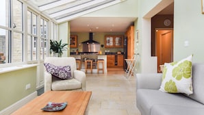 A cozy living room with a sofa, a chair, and a wooden coffee table. A kitchen is visible in the background with a dining area. - Barebones Farm - sleeps 14 guests  in 7 bedrooms (Winchcombe)